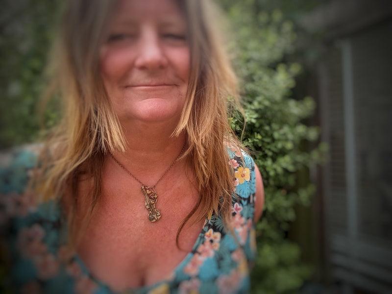 A close-up portrait of a woman outdoors wearing a floral-patterned teal top and a gold pendant necklace handmade by Elizabeth Shewan Jewellery Designer