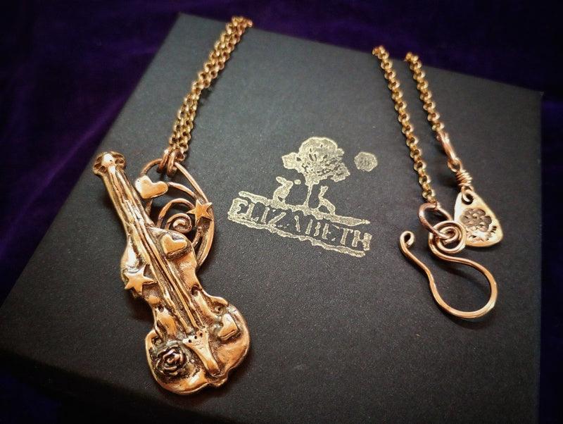 A gold-tone violin pendant necklace displayed on a black presentation box with the brand name Elizabeth. The necklace includes a detailed violin pendant, a matching chain, and a small heart-shaped charm.