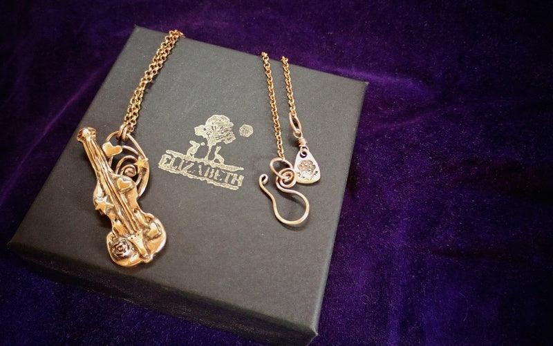 A gold-tone metal necklace with a violin-shaped pendant and a small heart-shaped charm, displayed on a dark gray gift box with the brand name 'ELIZABETH' printed on it, set against a purple velvet background.