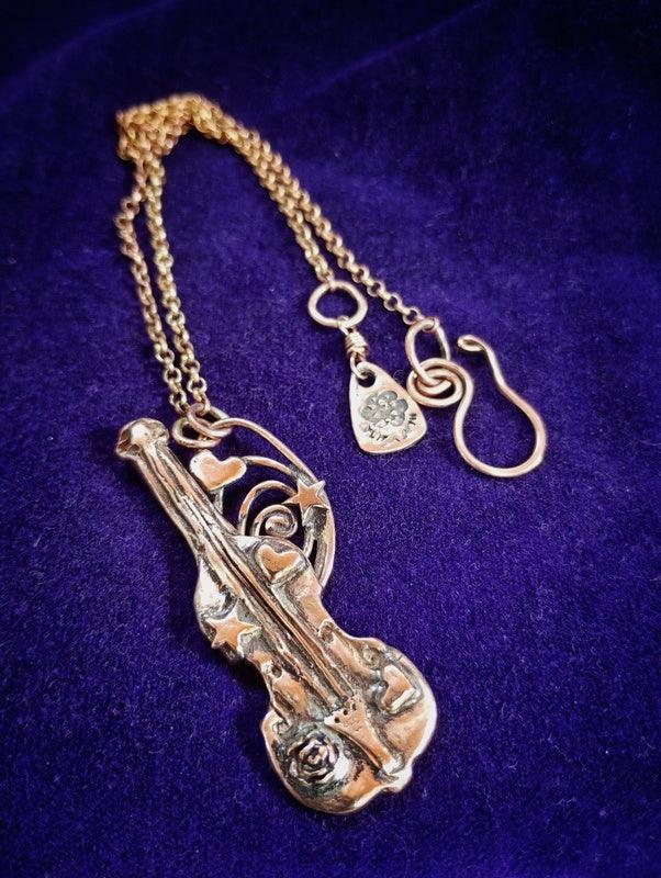 A gold-toned bronze violin pendant necklace with a matching chain and small charm, displayed on a textured dark purple velvet background by Elizabeth Shewan Jewellery Designer