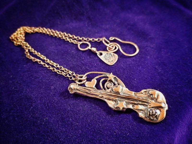 A gold-tone violin pendant necklace with a detailed violin charm and matching chain, displayed on a textured purple velvet background.