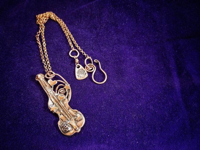 A gold-tone metal necklace featuring a violin-shaped pendant, a small hanging tag, and a treble clef charm, displayed on a textured purple velvet background by Elizabeth Shewan Jewellery Designer