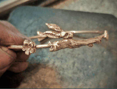 Rose gold bracelet with floral and branch design held by a hand on a textured surface.