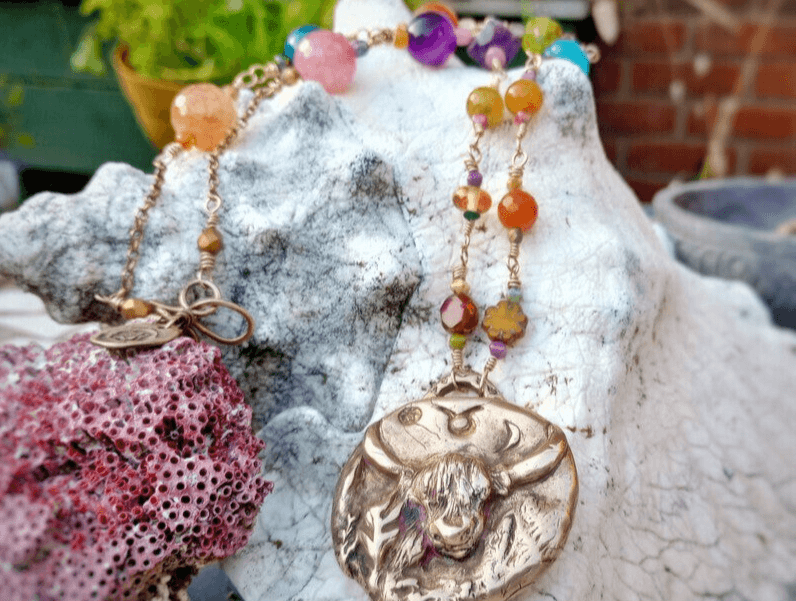 Zodiac Taurus bronze Pendant with a beaded crystal necklaces by Artist and Jewellery Designer Elizabeth Shewan on a textured stone surface, outside her studio in Dorset.