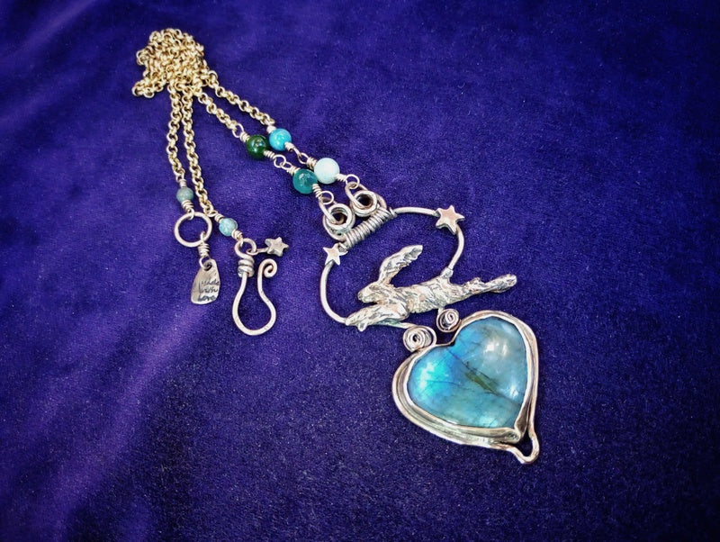 A handmade bohemian-style necklace featuring a large heart-shaped labradorite pendant, a silver crescent moon and rabbit charm, and turquoise beads on a gold chain, displayed on a purple velvet background.