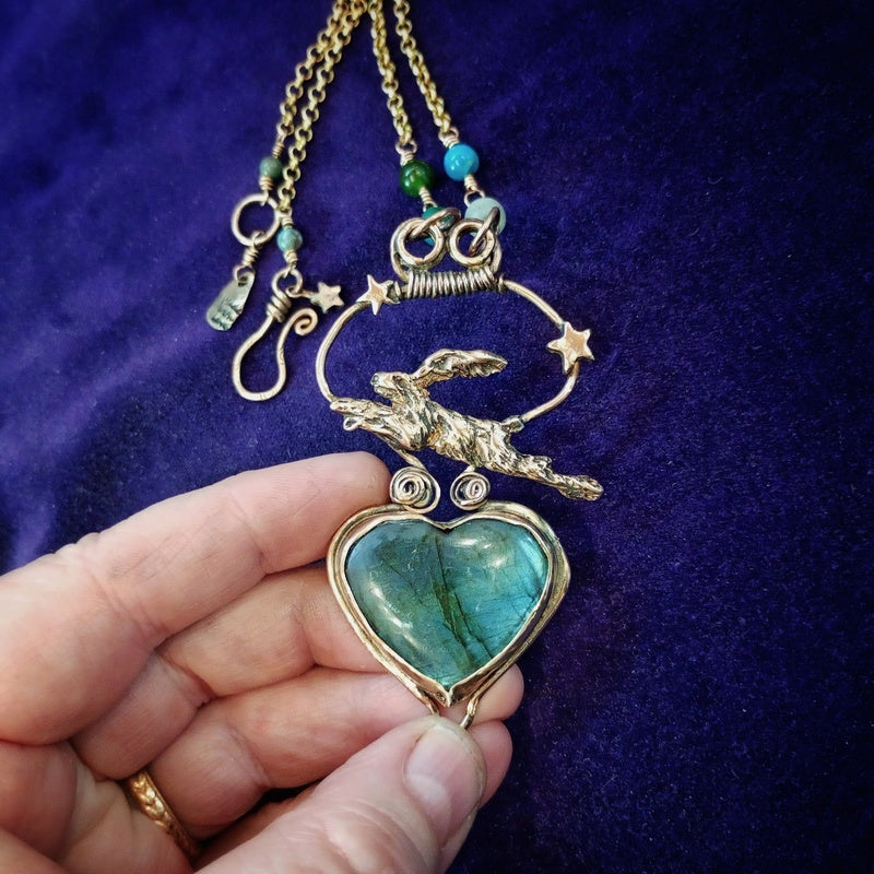 A gold-toned pendant necklace featuring a heart-shaped green stone pendant, a rabbit motif, stars, and turquoise beads, held by a hand against a dark purple velvet background.