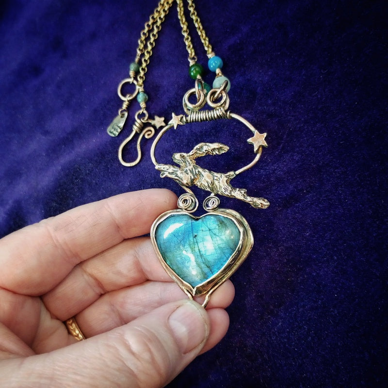 A close-up of a hand holding a gold-toned heart-shaped pendant with a blue-green labradorite stone, featuring a rabbit motif and crescent moon with stars on a dark purple velvet background.