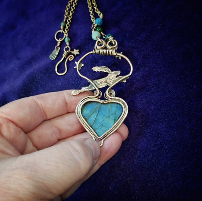 A hand holding a gold-toned necklace with a heart-shaped turquoise stone pendant, featuring a small eagle design above the heart, set against a dark purple velvet background.