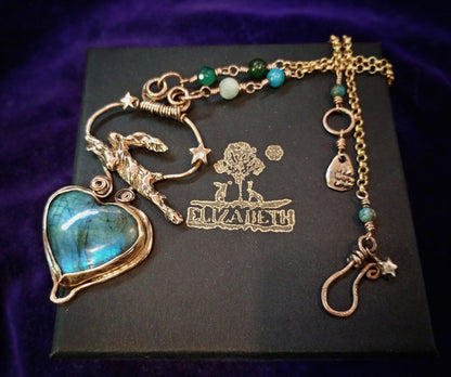 A handmade gold-toned pendant necklace featuring a large heart-shaped labradorite gemstone with blue-green iridescence, a crescent moon with stars, a deer charm, and a skull charm, displayed on a black box with the name 'ELIZABETH' engraved, set against a dark purple velvet background.