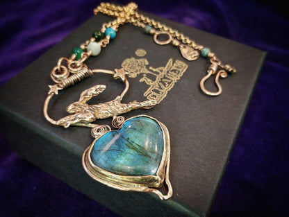 A gold-toned pendant necklace featuring a heart-shaped labradorite stone with blue-green iridescence, displayed on a black box over a purple velvet background.