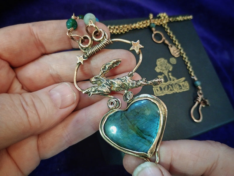 A handmade gold-toned pendant necklace featuring a large heart-shaped labradorite stone with blue-green iridescence, a detailed rabbit figurine, and star accents, held in a hand against a dark purple velvet background with a black box.