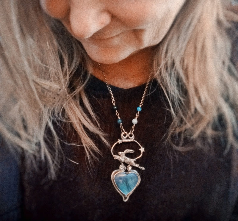 A close-up lifestyle shot of a woman wearing a silver-toned bird pendant necklace with a light blue heart-shaped stone and small turquoise beads on the chain.