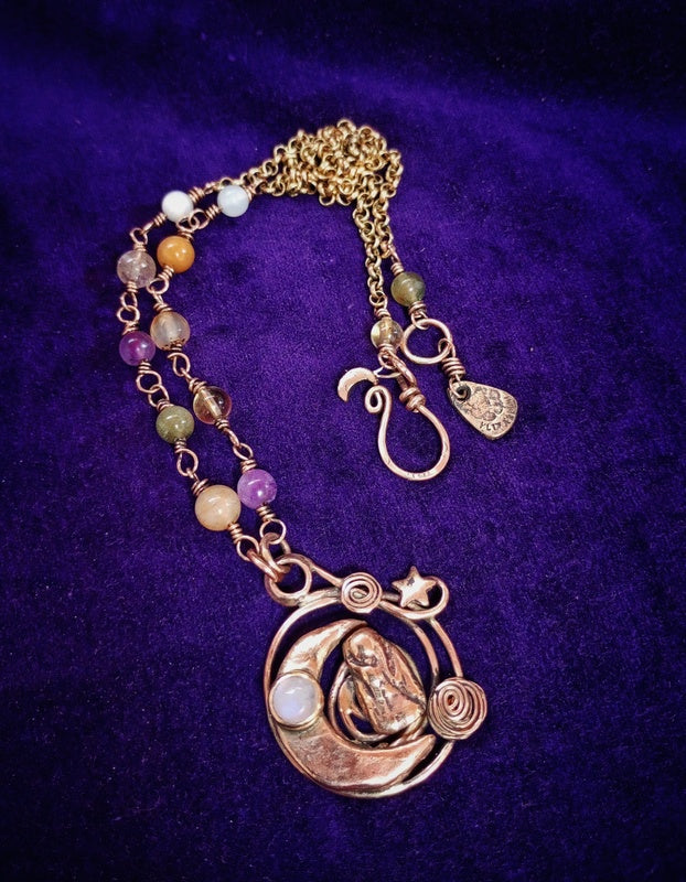 A handmade bohemian-style necklace featuring a gold-toned crescent moon and star pendant with a small figurine, accented by multi-colored gemstone beads and a heart charm, displayed on a deep purple velvet background.