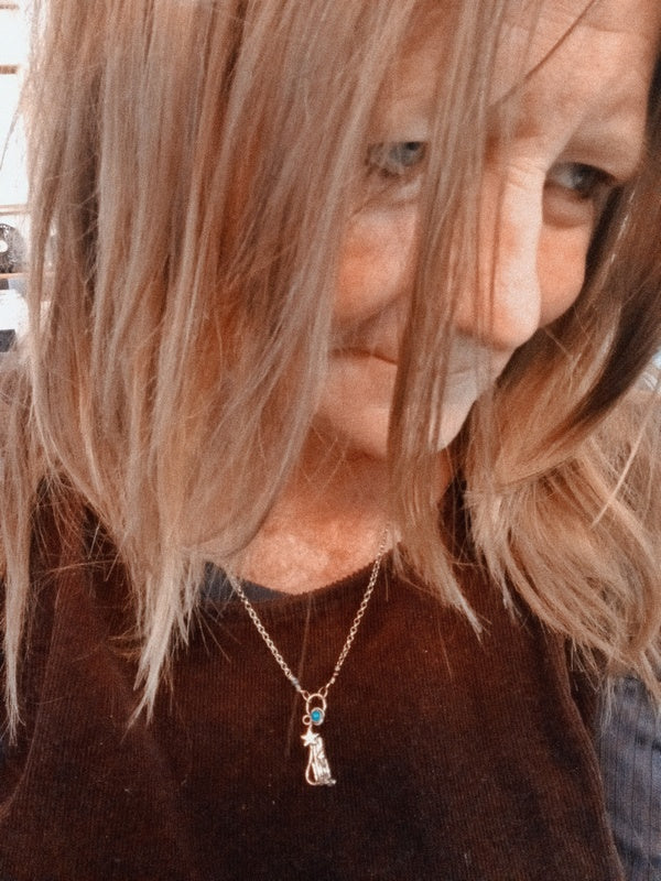 A close-up lifestyle shot of a woman with shoulder-length blonde hair wearing a delicate silver pendant necklace with a small cat charm and a tiny blue stone accent.