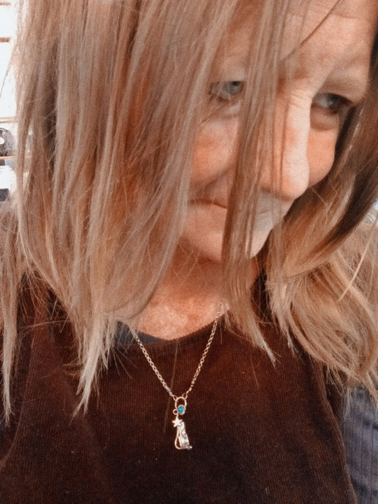 A close-up lifestyle shot of a woman with shoulder-length blonde hair wearing a delicate silver pendant necklace with a small cat charm and a tiny blue stone accent.