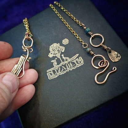 A close-up of a hand holding a gold-toned pendant necklace next to a black presentation box with a gold 'Elizabeth' tree logo. The necklace features a delicate chain, a circular ring, a spiral charm, a small textured tag, and a green bead accent.