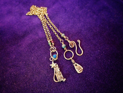 A gold-toned triple-strand chain necklace with celestial charms, including a star, crescent moon, and a small bag charm, accented with turquoise beads, displayed on a textured purple background.