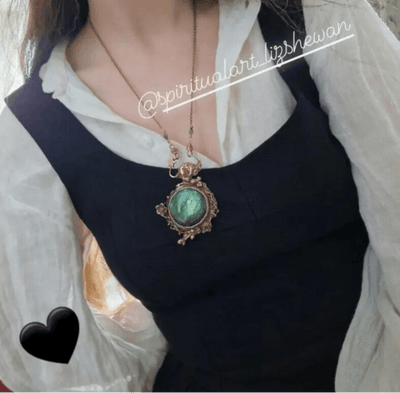 Person wearing a black apron over a white shirt with a decorative bronze handmade necklace by Elizabeth Shewan featuring a green labradorite crystal and hare in nature undergrowth.
