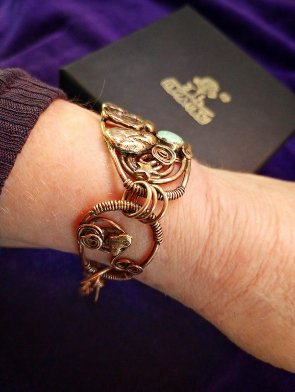 A close-up of a person's wrist wearing a copper-toned metal cuff bracelet with floral and leaf motifs, a small light green stone, and a frog detail. A black branded box is visible in the background on a purple fabric surface.