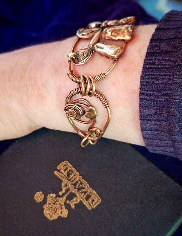 A handmade copper wire bracelet with organic, organic-shaped links and coiled wire details, shown worn on a person's wrist. A dark purple knit cuff is visible, and a dark box with a gold foil logo is in the foreground.