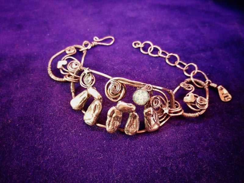 A handmade gold-toned wire bracelet featuring textured stone charms and spiral wire details, displayed on a textured purple background. Stonehenge Bracelet.