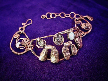 Stonehenge Bracelet. A handmade copper wire bracelet featuring textured stone-like elements, spiral wire details, and a small light blue stone, displayed on a purple velvet background.