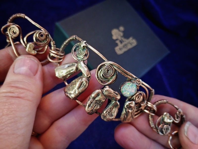A handmade gold-toned bracelet held in two hands, featuring natural opal stones, textured gold beads, and coiled wire details. A dark blue velvet box with a gold logo is visible in the background.