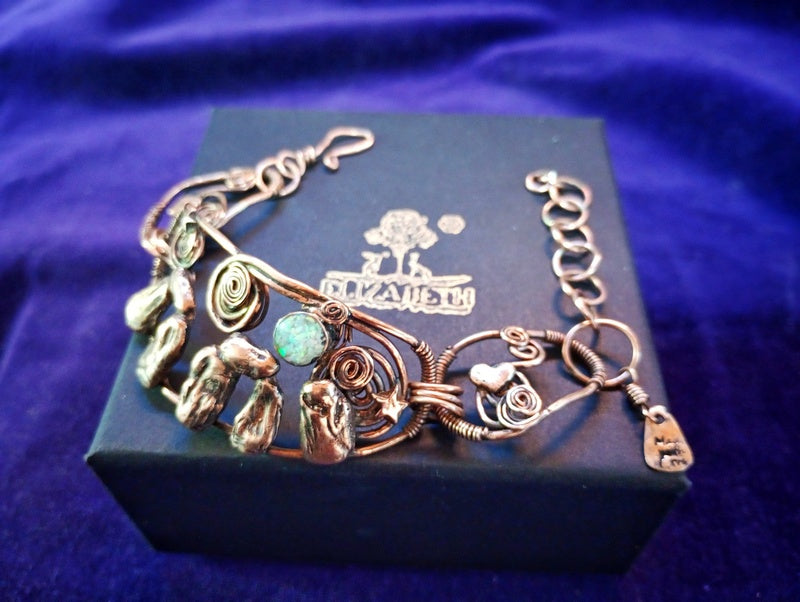 A handmade copper wire bracelet with turquoise and green stone accents, displayed on a dark blue jewelry box with the brand name 'El Mariachi' visible on the lid, set against a purple velvet background. Stonehenge Bracelet. Stonehenge Bracelet.