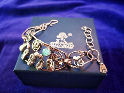 A handmade copper wire bracelet with turquoise and green stone accents, displayed on a dark blue jewelry box with the brand name 'El Mariachi' visible on the lid, set against a purple velvet background. Stonehenge Bracelet. Stonehenge Bracelet.
