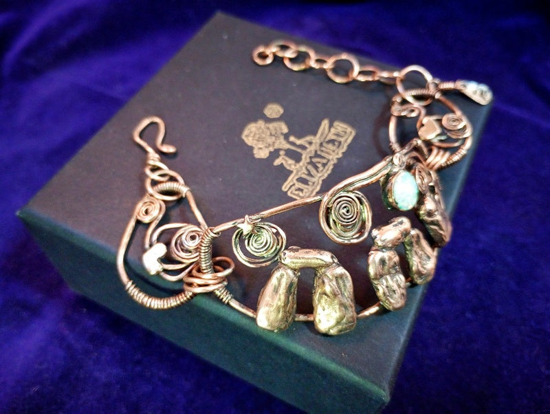 A handmade wire-wrapped bracelet with natural stone accents and spiral details, displayed on a dark green jewelry box against a deep purple velvet background. Stonehenge Bracelet.