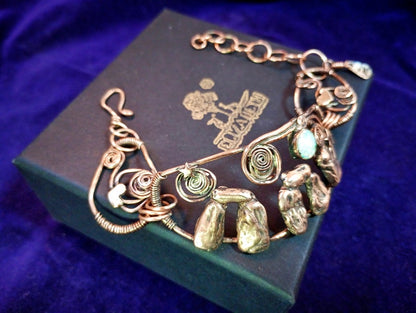 A handmade wire-wrapped bracelet with natural stone accents and spiral details, displayed on a dark green jewelry box against a deep purple velvet background. Stonehenge Bracelet.