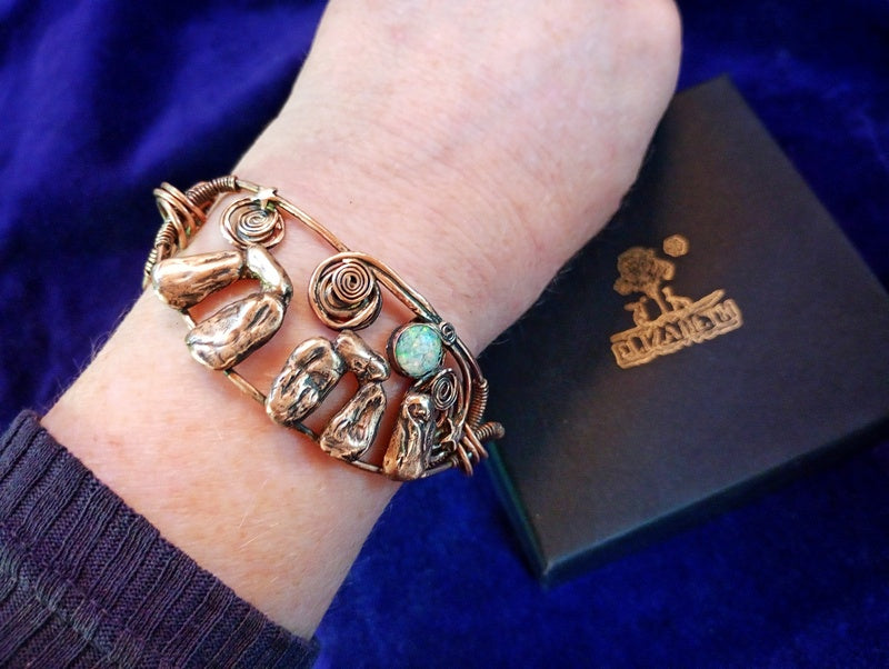 A close-up of a person's wrist wearing a handmade copper-toned wire bracelet with sculpted animal and organic shapes, a spiral detail, and a small turquoise stone. A dark brown jewelry box with a gold logo is visible in the background against a deep blue velvet surface. Stonehenge Bracelet.