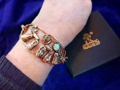 A close-up of a person's wrist wearing a handmade copper-toned wire bracelet with sculpted animal and organic shapes, a spiral detail, and a small turquoise stone. A dark brown jewelry box with a gold logo is visible in the background against a deep blue velvet surface. Stonehenge Bracelet.