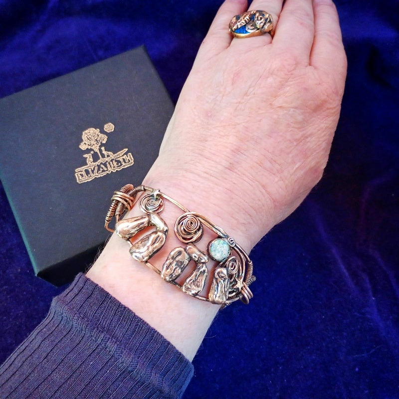A close-up of a person's wrist wearing a gold-toned multi-piece jewelry set, including a cuff bracelet with sculpted animal figures and a ring, against a dark blue velvet background with a black branded box. Stonehenge Bracelet.