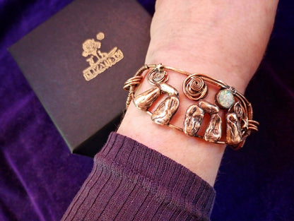 Stonehenge Bracelet. A close-up of a person's wrist wearing a copper-toned cuff bracelet with textured stone-like details and a small round light stone. A dark purple velvet fabric and a dark box with a gold logo are visible in the background.