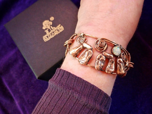 Stonehenge Bracelet. A close-up of a person's wrist wearing a copper-toned cuff bracelet with textured stone-like details and a small round light stone. A dark purple velvet fabric and a dark box with a gold logo are visible in the background.