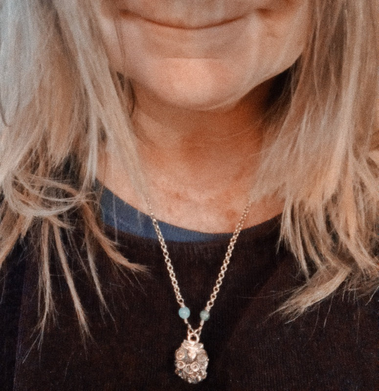 A close-up of a person wearing a delicate gold chain necklace with a textured, clear crystal pendant and small light blue beads, paired with a dark brown knit top.