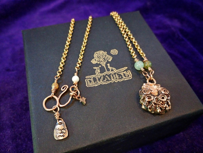 A pair of gold-toned jewelry pieces displayed on a black box with the brand name 'ELIZABETH'. The left piece features a curly ring clasp with a small hanging charm, and the right piece has an ornate sheep head pendant with a light green bead accent.