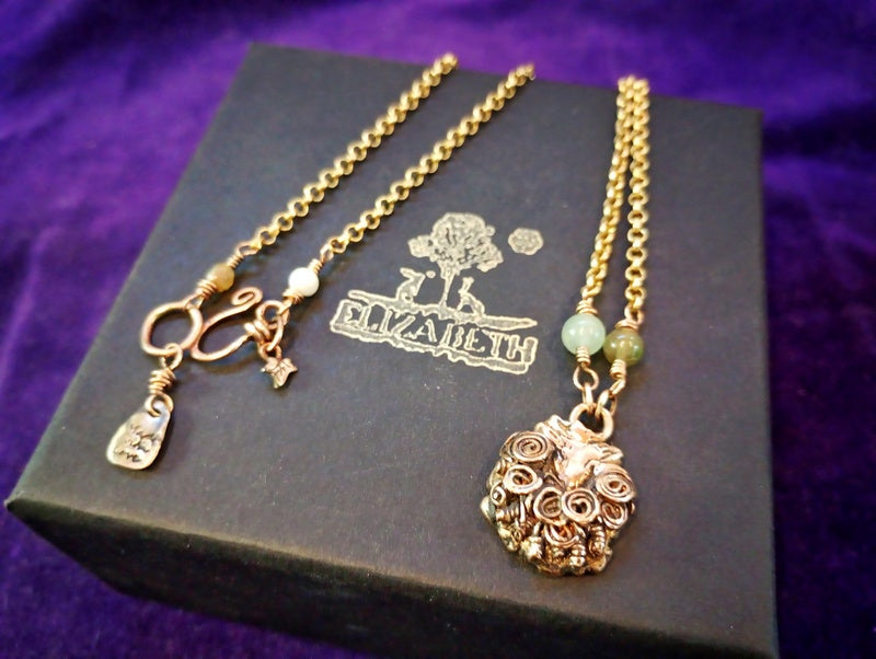 A gold-toned jewelry set displayed on a black branded box against a purple fabric background. The set includes a chain necklace with a floral pendant, a bracelet with a toggle clasp and charm, and a small green bead accent.