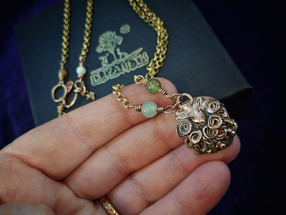 A close-up of a hand holding a gold-toned necklace with a detailed spiral pendant and two light green beads, with a dark branded box in the background.