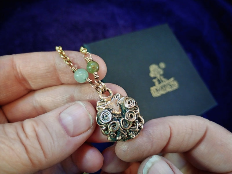 A close-up of a hand holding a gold-toned pendant necklace with green beads, with a dark branded box in the background.