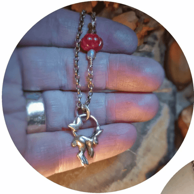Necklace with a red bead and silver charm held between fingers against a blurred natural background